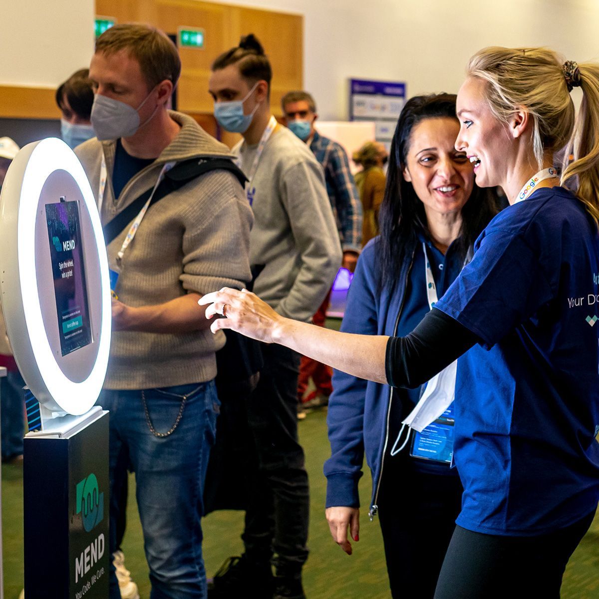 An attendee interacts with a ring light photo booth while a staff member in a blue shirt assists in an indoor space.