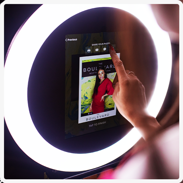 A person uses their finger to share a photo from a digital photo booth featuring a glowing ring light.
