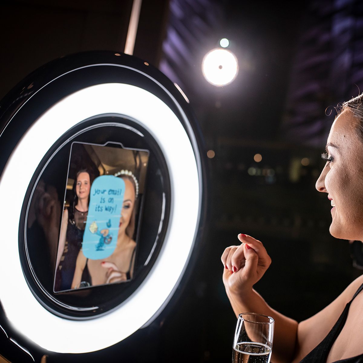 A person interacts with a circular ring light photo booth, smiling as a screen displays a message about a photo delivery.