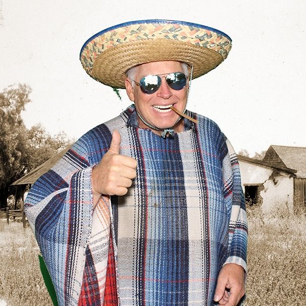 A smiling person in a sombrero and blue plaid poncho gives a thumbs-up while holding a cigar in a rustic outdoor setting.
