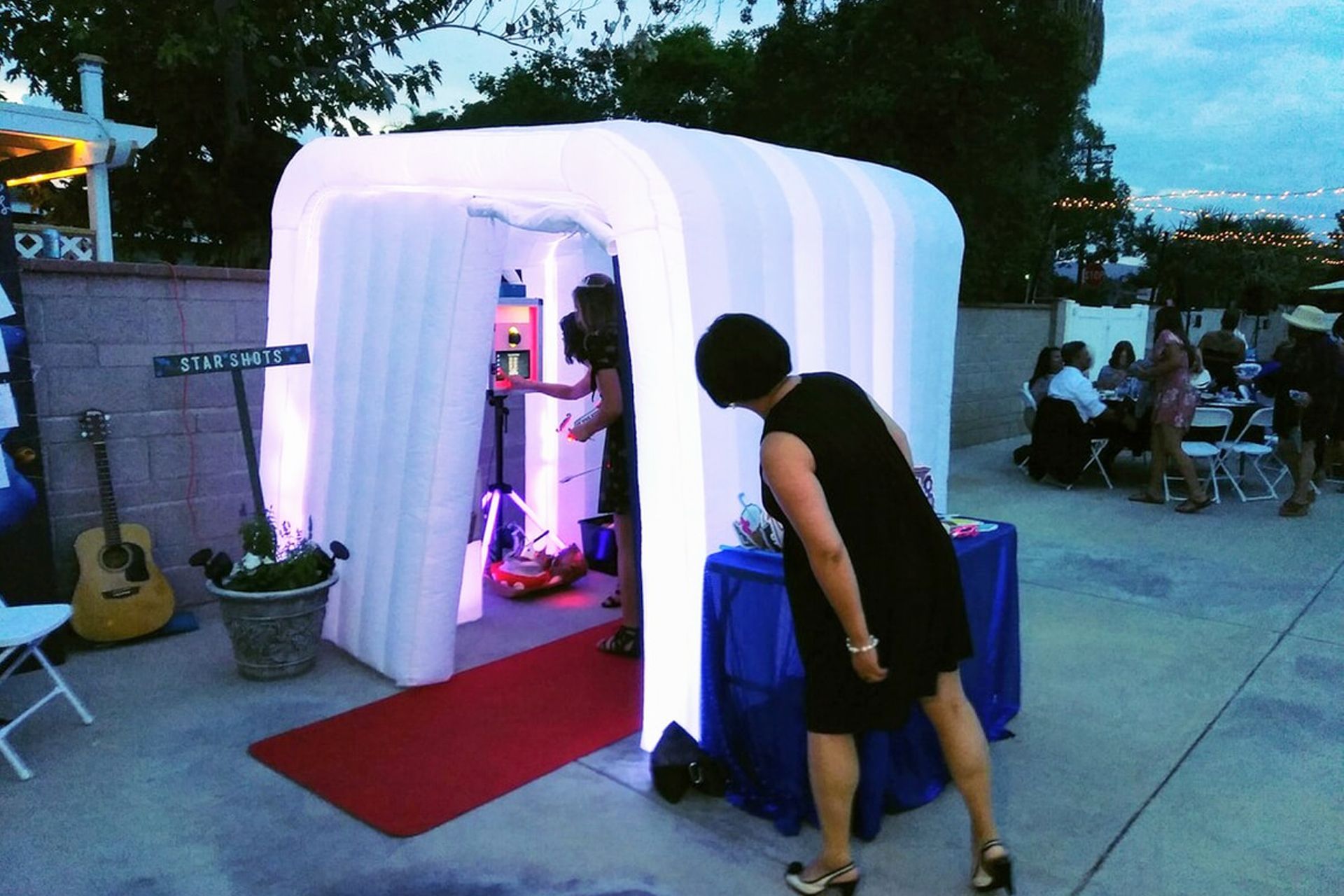 A white, inflatable, glowing photo booth enclosure stands on a patio with a red carpet runner and people nearby.