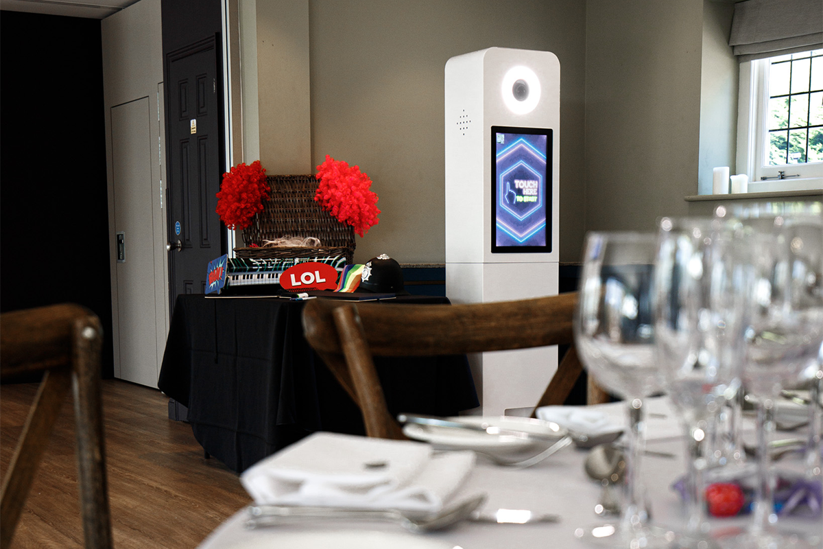 A white photo booth stands in a room behind a dining table with place settings, near a table decorated with red pom-poms.