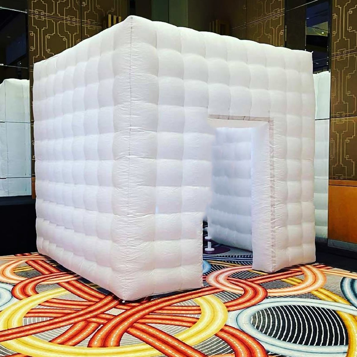 A white, inflatable, cubical photo booth enclosure sits on a patterned carpet inside a room.