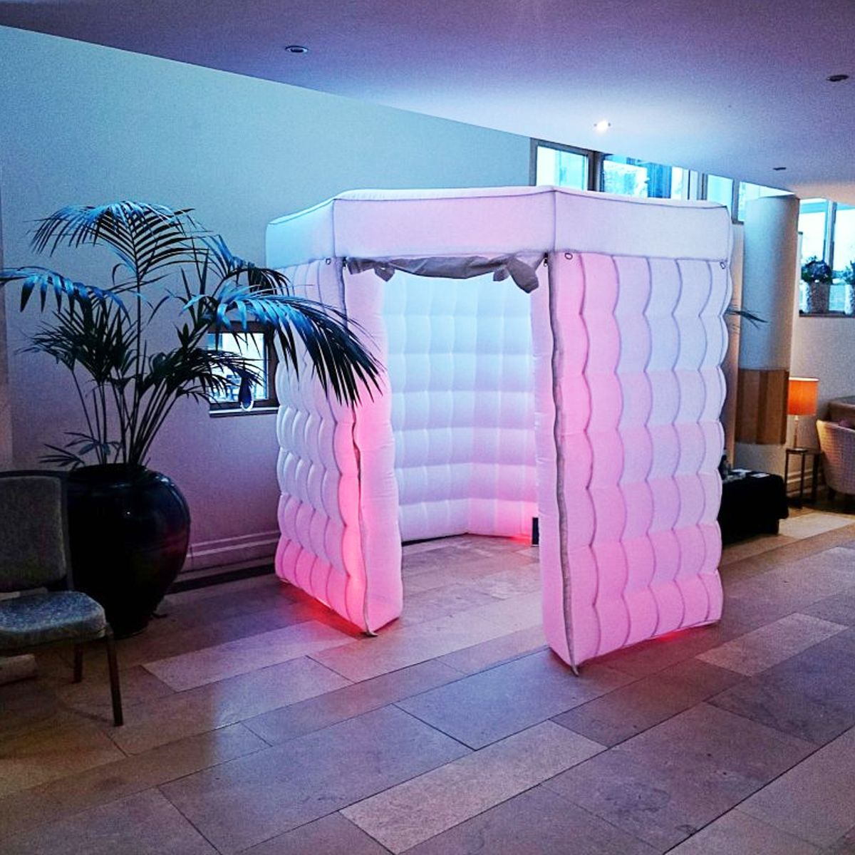An illuminated, inflatable, modular booth with pink interior lighting stands on a stone floor next to a large potted plant.