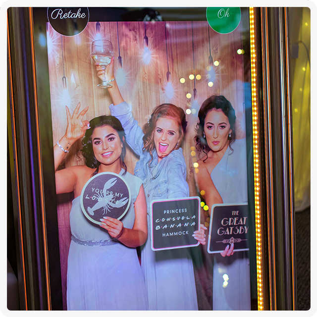 Three people pose for a photo in a mirror photo booth, holding props with text while smiling against a light-bulb backdrop.