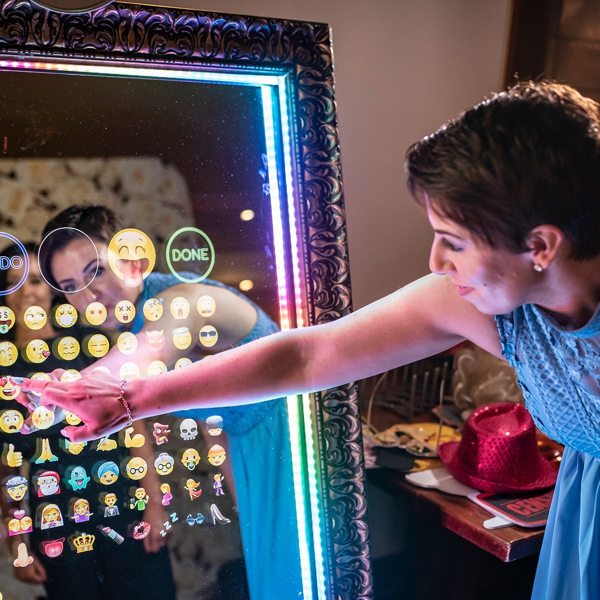 A person in a blue dress interacts with a touch-screen mirror display filled with emojis at an event.