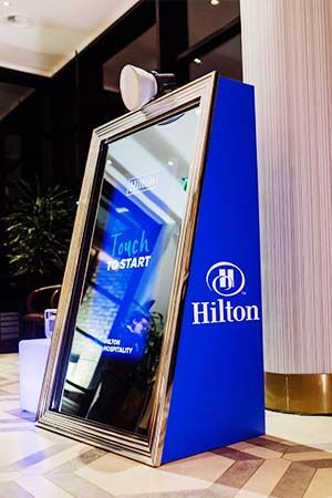 A vertical, blue, Hilton-branded interactive mirror photo booth standing in an indoor hotel lobby.