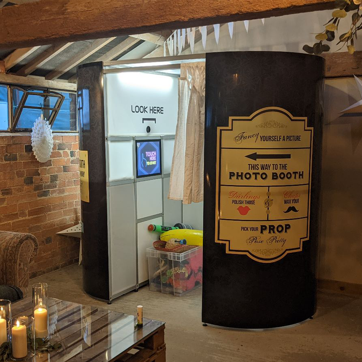 A portable photo booth with an open entrance and a sign pointing to props, set up in a rustic, brick-walled indoor space.