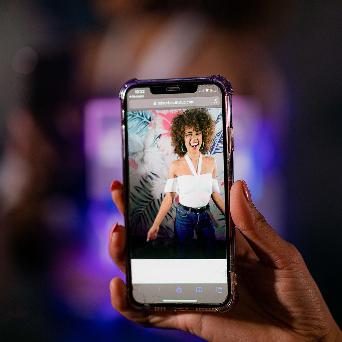 A hand holds a smartphone displaying a photograph of a smiling person with curly hair wearing a white top and blue jeans.
