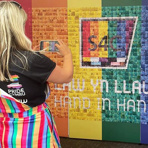 A person in a rainbow outfit takes a photo of an S4C pride-themed art installation with a rainbow background.