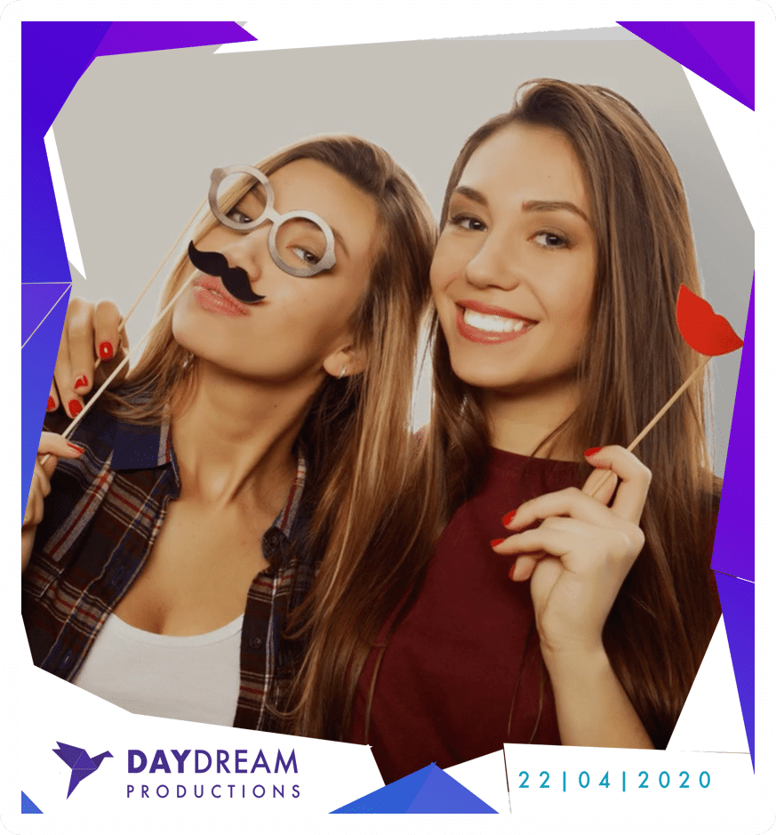 Two people pose in a photo booth, one wearing mustache and glasses props, the other holding a red lip prop.