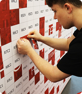 A person carefully placing a square sticker labeled R6C4 onto a grid wall patterned with red and white squares.