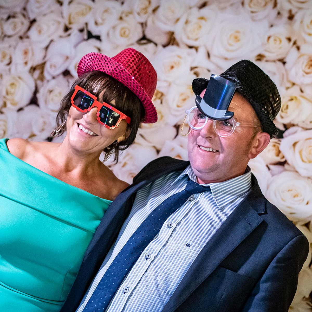 A smiling couple posing in front of a white floral backdrop, wearing festive props including hats and oversized glasses.