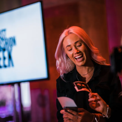 A person with light-colored hair smiles while holding a small photograph and a smartphone, set against a blurry screen.