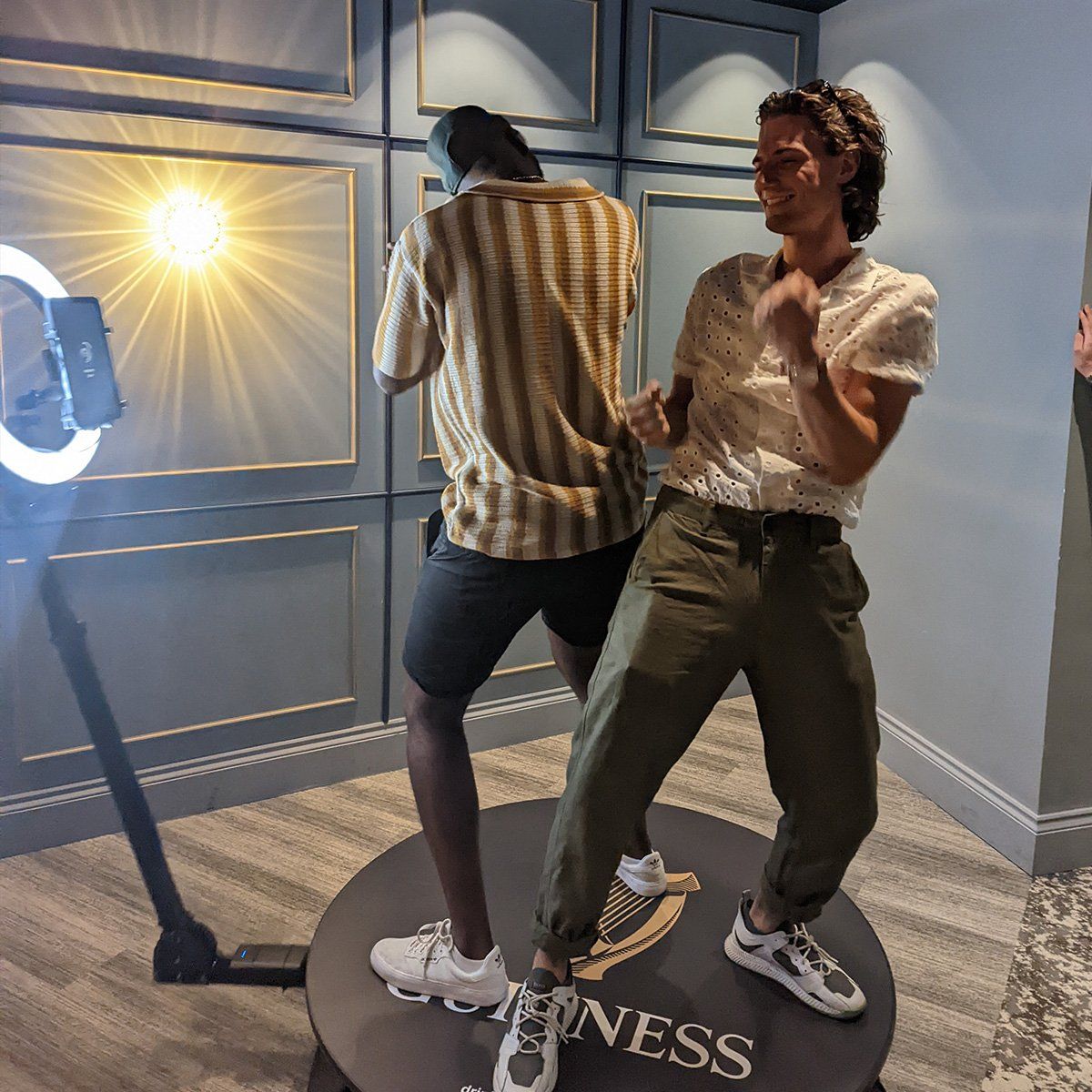 Two people dance on a rotating circular platform in front of a ring light and a paneled wall with a Guinness logo.