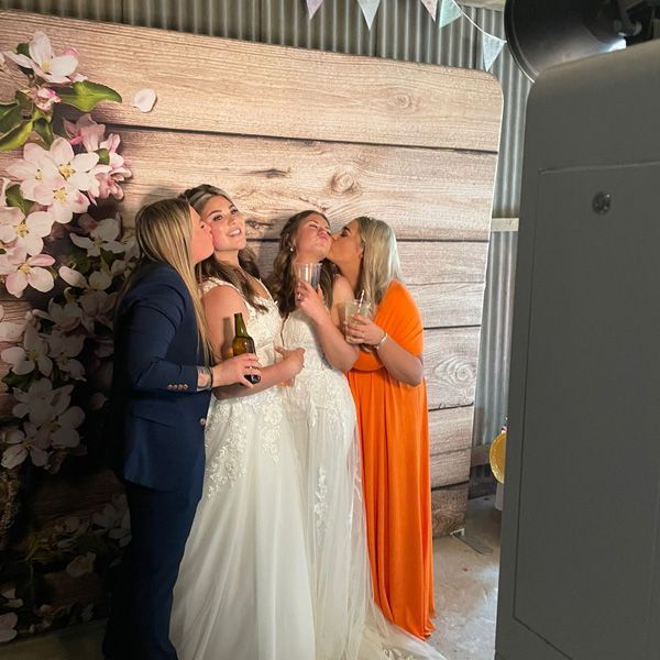 Two people in white wedding dresses stand between two others in suits and gowns, all posing for a photo at a party.