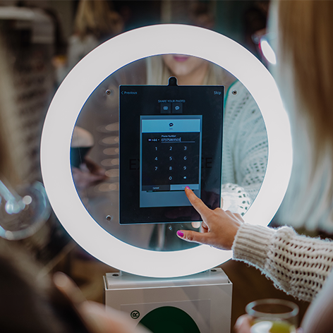 A person touches a touchscreen keypad on a portable photo booth ring light set up at an indoor event.