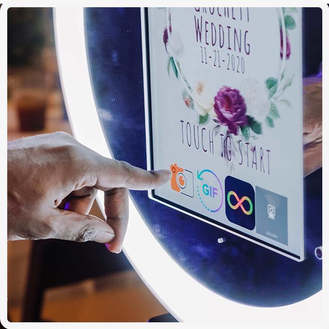 A hand touching a touch-screen photo booth kiosk at a wedding, with options to start a photo, GIF, or Boomerang.