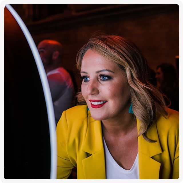 A person in a yellow blazer smiling while looking at a circular light feature in a dimly lit room.