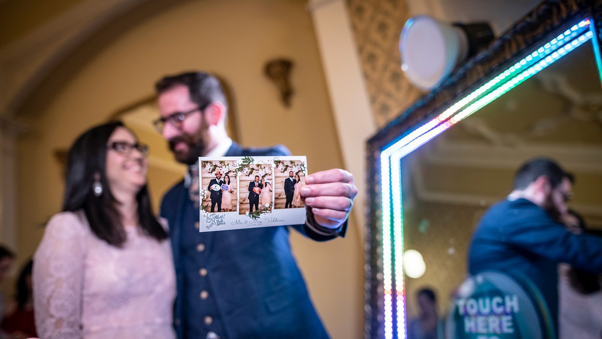 A smiling couple in formal attire shows a photo strip taken at a mirror-style event photo booth.