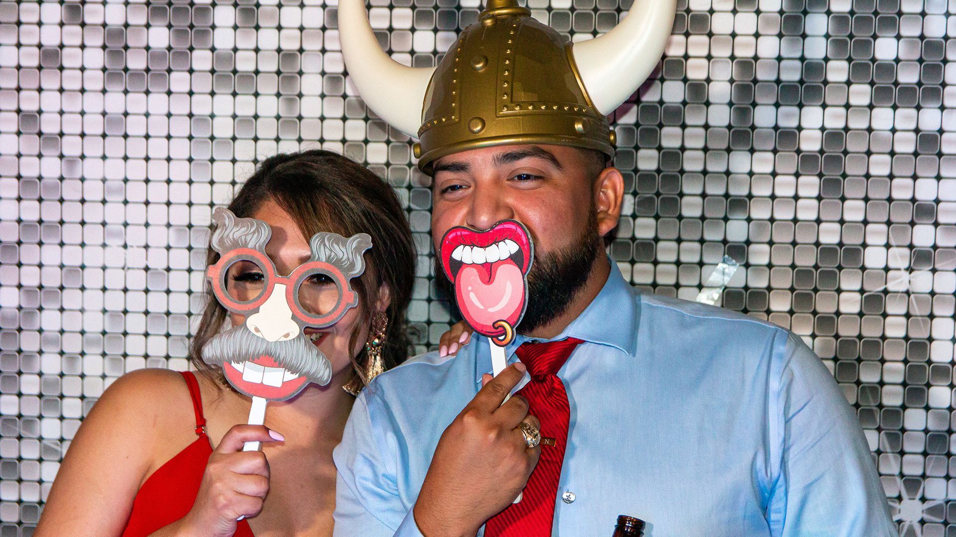 Two people pose in front of a silver sequined backdrop, holding up photo booth props of large glasses and a mouth.