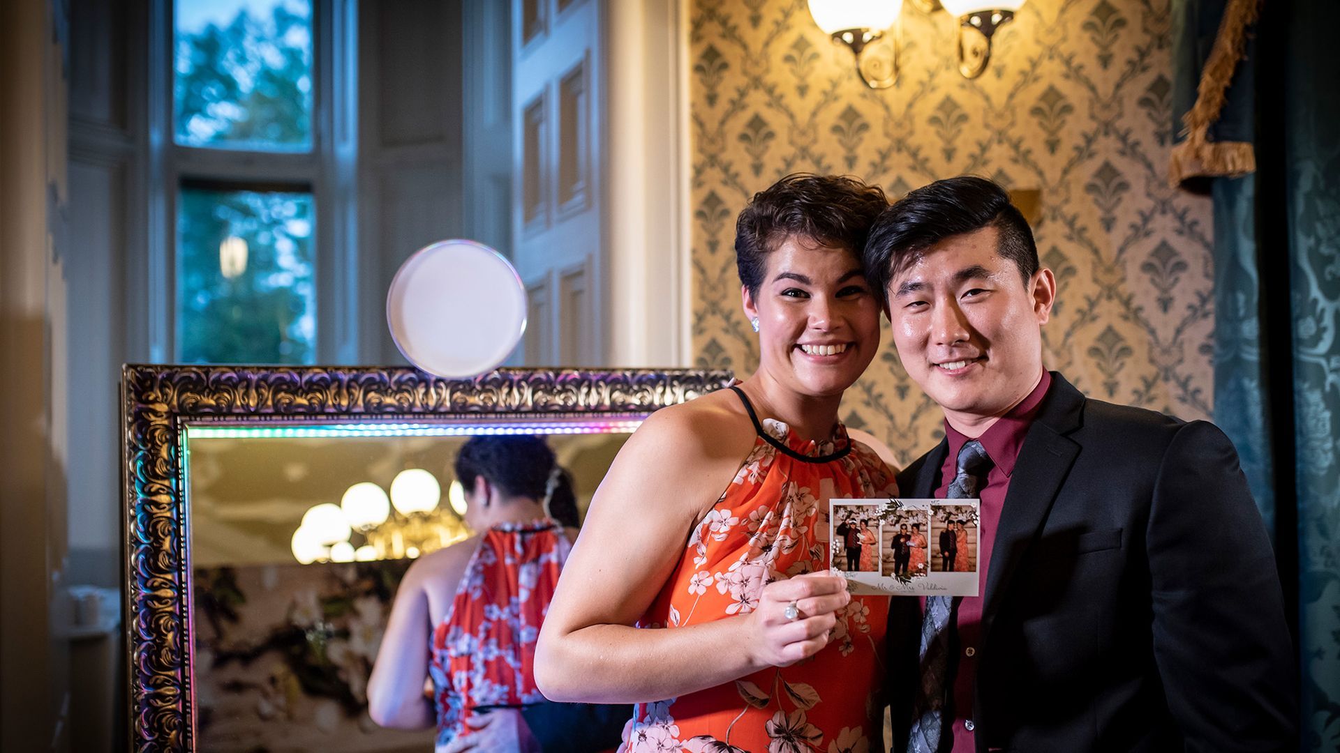 A smiling couple holds a small photo strip in front of a lit mirror in a room with patterned wallpaper and a large window.