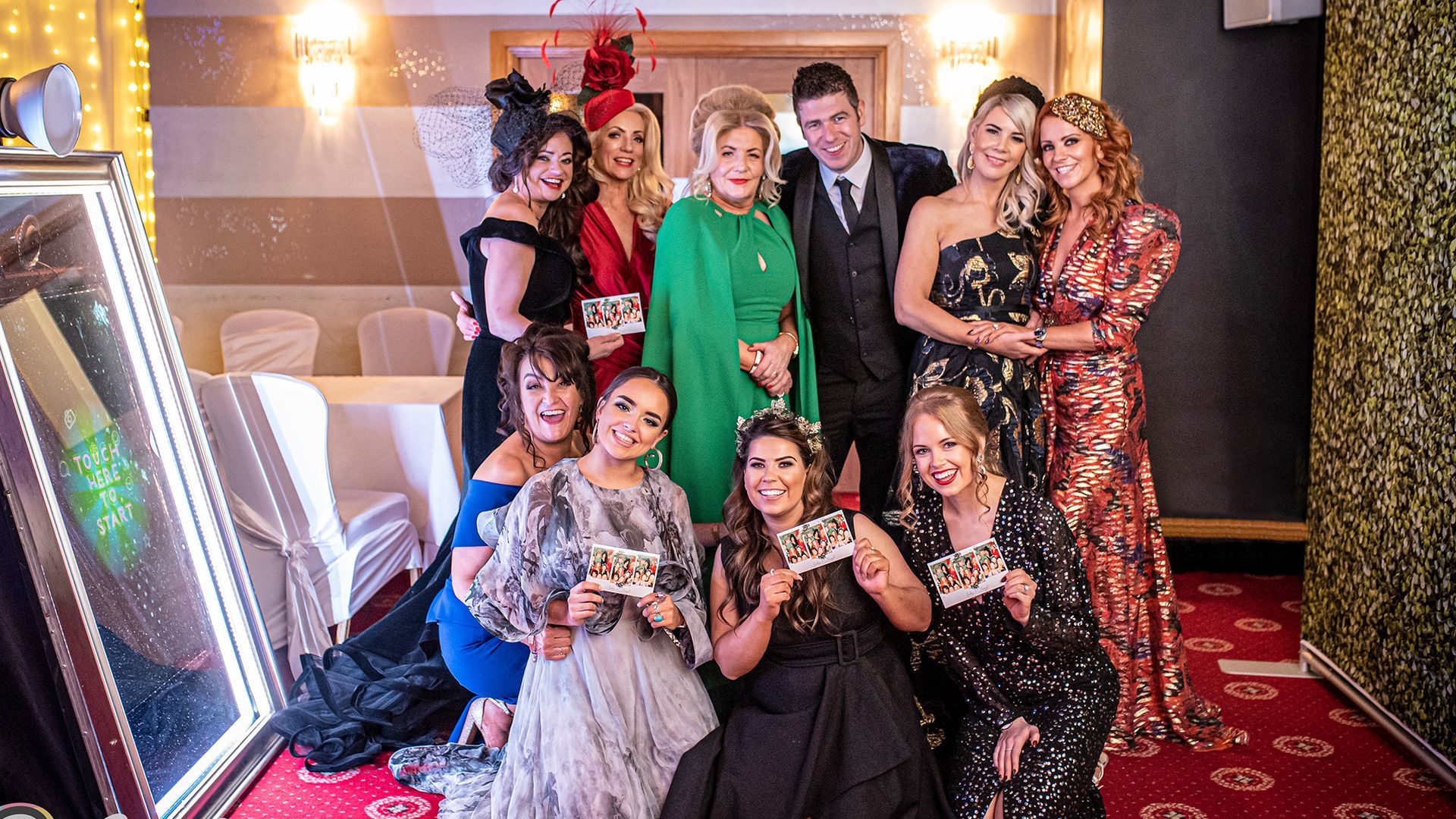 A group of people in formal attire pose for a photo at an event, with some holding small props in front of a mirror.