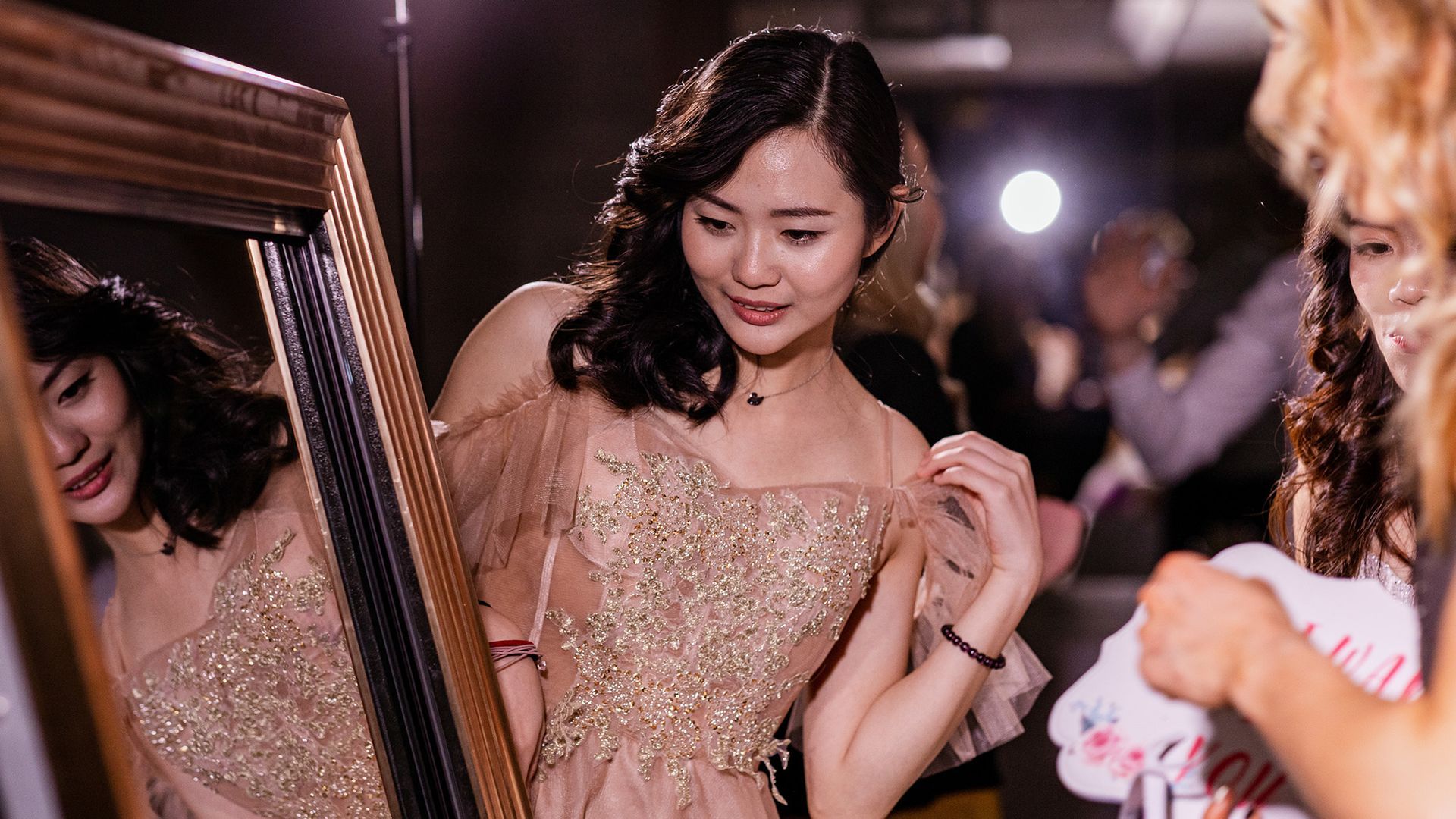A person in a beaded, light-colored formal gown adjusts their dress while looking at their reflection in a mirror.