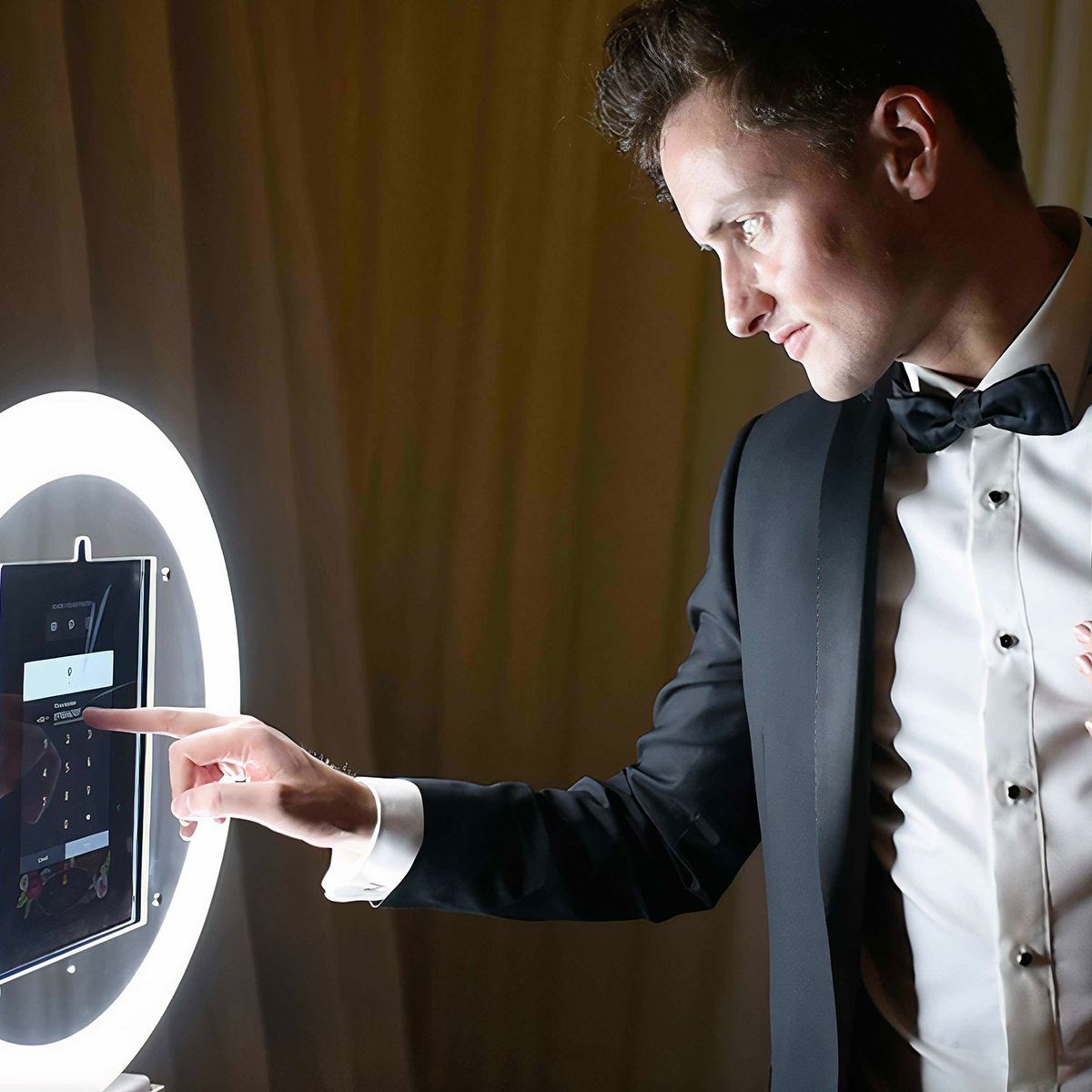 A person in a tuxedo uses their finger to touch a screen on a ring light photo booth.