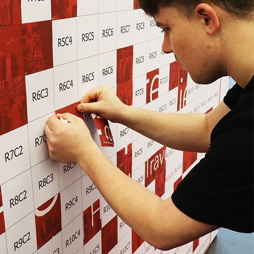A person attaches a square tile with a grid label to a wall covered in a pattern of red and white numbered labels.