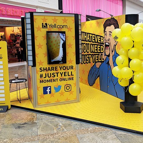 A Yell.com promotional kiosk in a mall featuring yellow branding, social media icons, and a graphic of a shouting person.