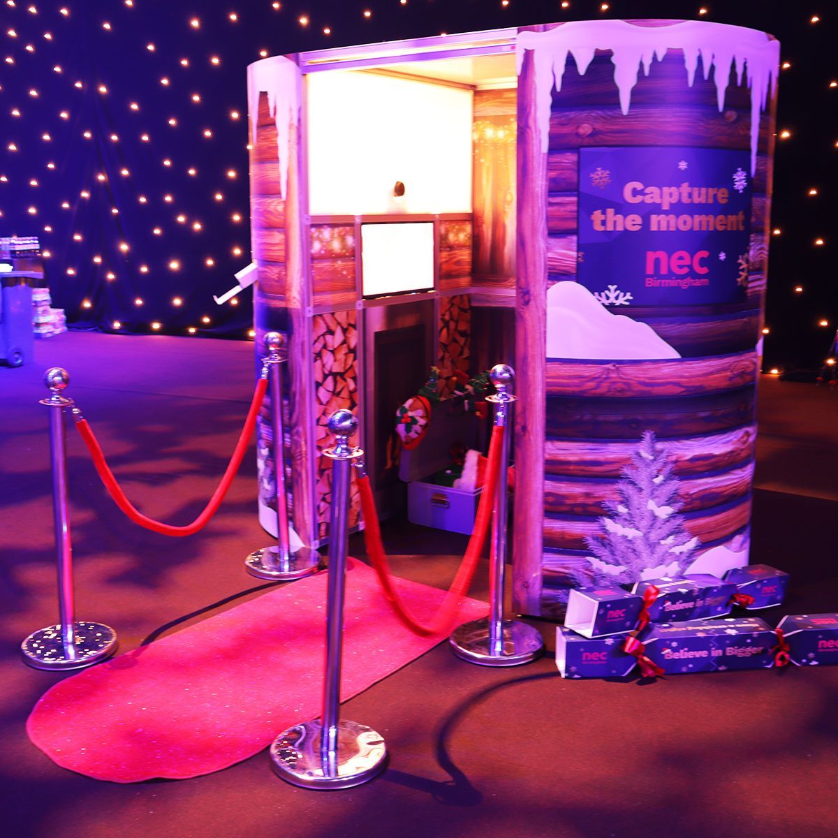 A winter-themed photobooth with log cabin graphics, a red velvet rope stanchion, and a small Christmas tree.