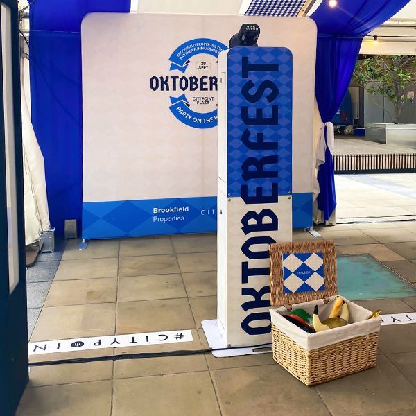 An Oktoberfest photo booth set up with a branded backdrop, a vertical sign, blue curtains, and a prop basket.