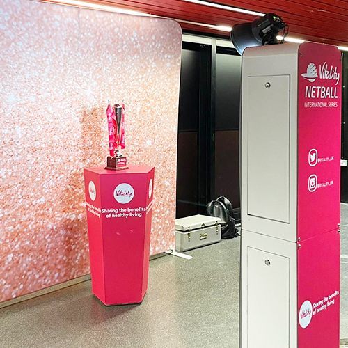 A trophy sits on a pink, branded plinth in front of a glittery backdrop, next to a Vitality Netball promotional kiosk.
