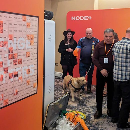People gather near a Node4 branded backdrop with a golden retriever and a photo booth.