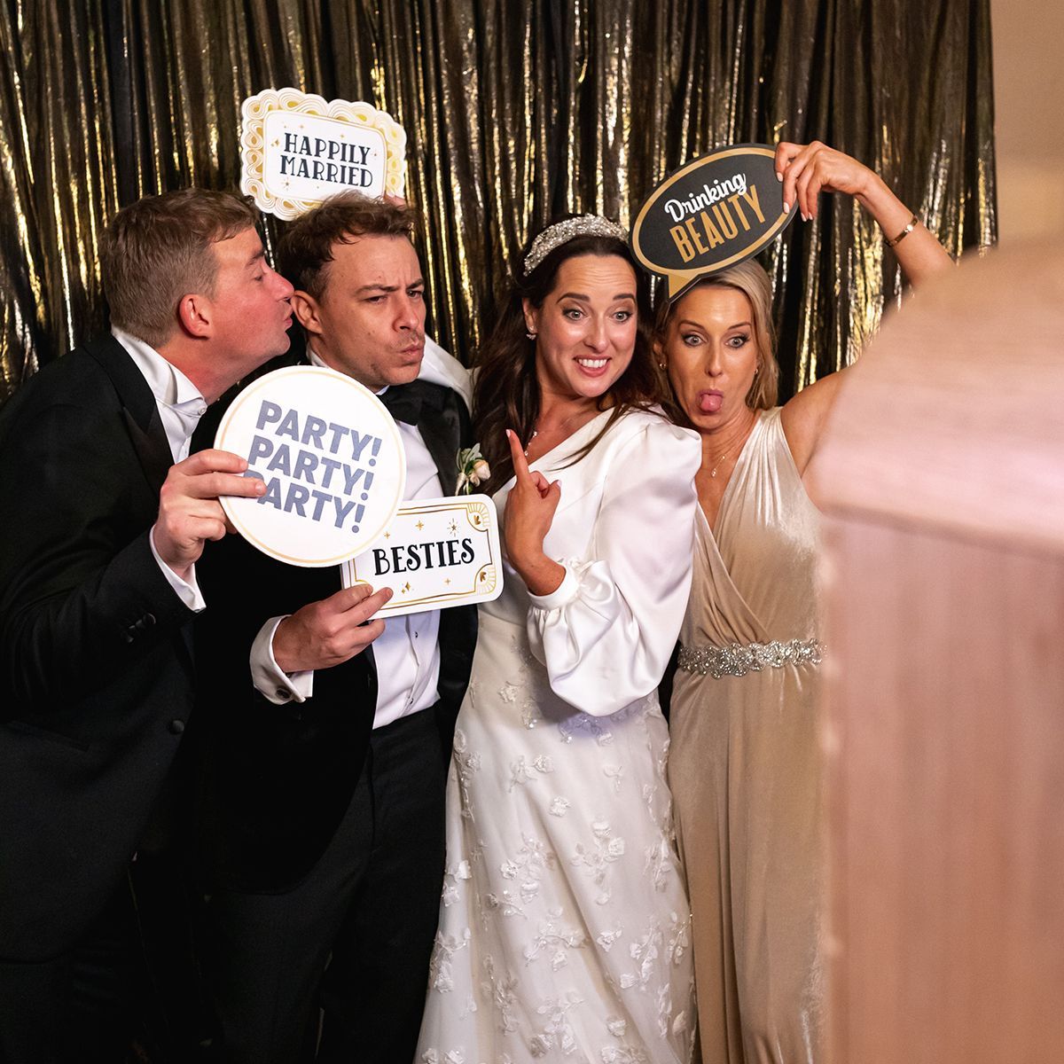 Four people in a photo booth pose with fun props against a gold backdrop, smiling and making playful expressions.