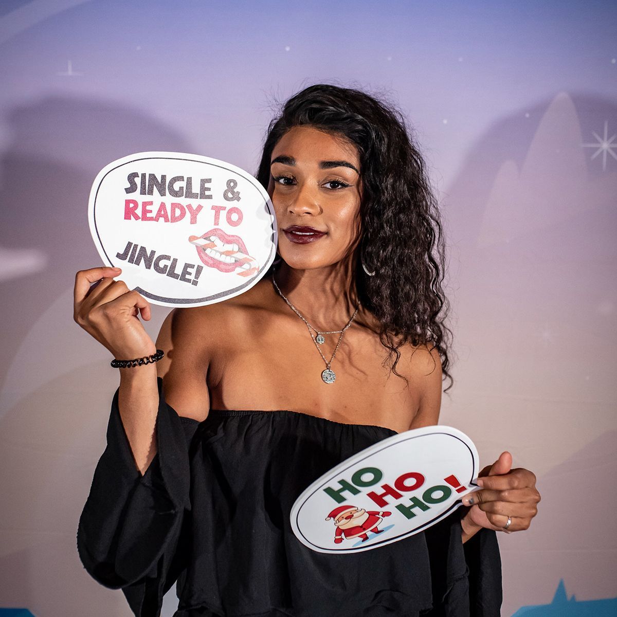 A person with dark curly hair wearing a black off-the-shoulder top holds signs saying