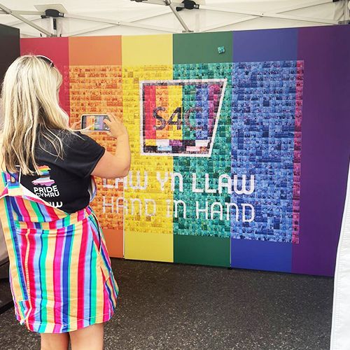 A person wearing a rainbow striped apron photographs a large, colorful display wall featuring the text