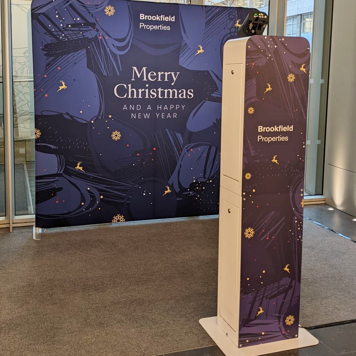 A navy blue holiday photo booth backdrop with festive text next to a matching branded kiosk in a bright indoor lobby.