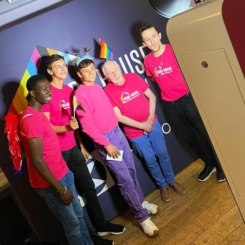 Five people in pink Pride House shirts pose together in front of a dark blue wall with a rainbow graphic.