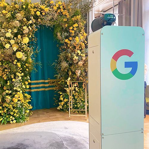 A large Google logo on a light blue kiosk stands next to a lush, yellow floral archway with a teal fabric backdrop.