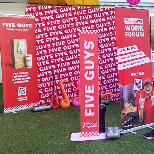 Five Guys recruiting display with red banners on artificial grass featuring a food photo and QR code for job applications.