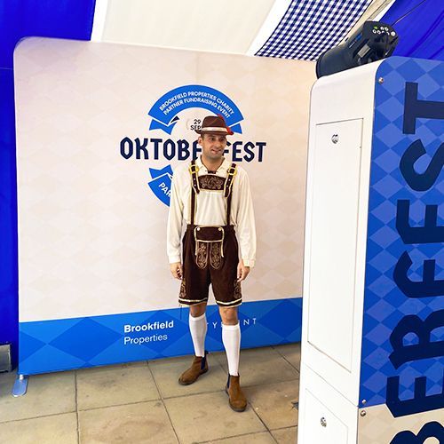 A person wearing traditional Bavarian lederhosen stands in front of a blue and white Oktoberfest event backdrop.