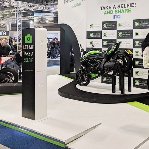 A green and black motorcycle on display at a trade show next to a selfie station kiosk with branded event signage.
