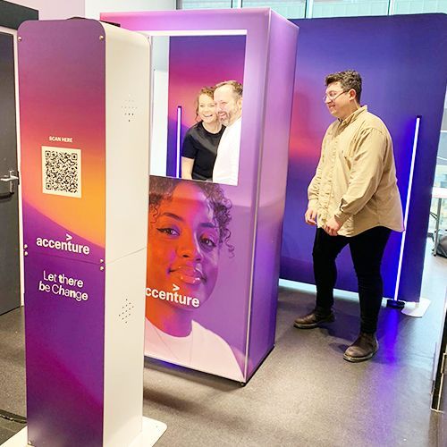 A person walks past an Accenture photo booth featuring a QR code, branded signage, and a person’s portrait.