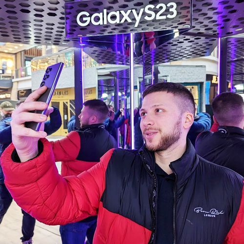 A person in a red and black puffer jacket takes a selfie with a Samsung Galaxy S23 in a brightly lit indoor mall display.