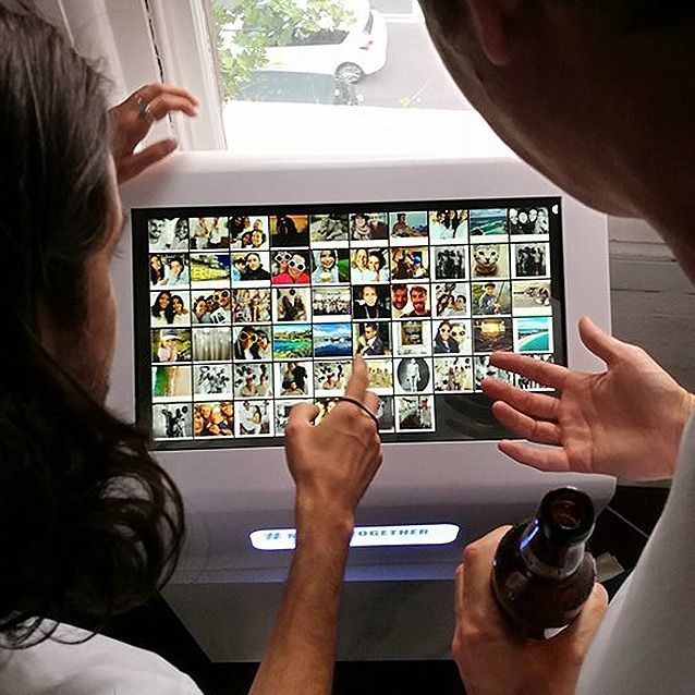 Two people interact with a touchscreen kiosk displaying a grid of photos, one person pointing at a specific image.