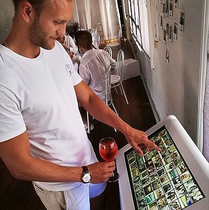 A person in a white shirt holds a cocktail while interacting with a digital touch-screen photo display at an event.