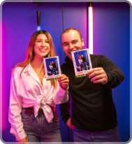 A smiling couple holds up matching photo booth prints in front of a blue wall with vibrant pink and blue neon lights.