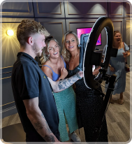 A group of people gathers around a ring light in a room with paneled walls, laughing while using a photo booth device.